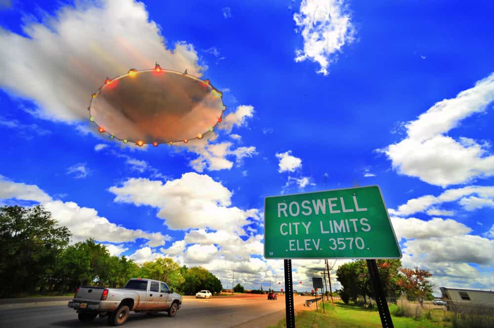 A large, illuminated UFO hovers in a partly cloudy blue sky above a Roswell city limits sign and cars on a road, inviting curiosity seekers and fans to explore with the ultimate Roswell vacation guide.