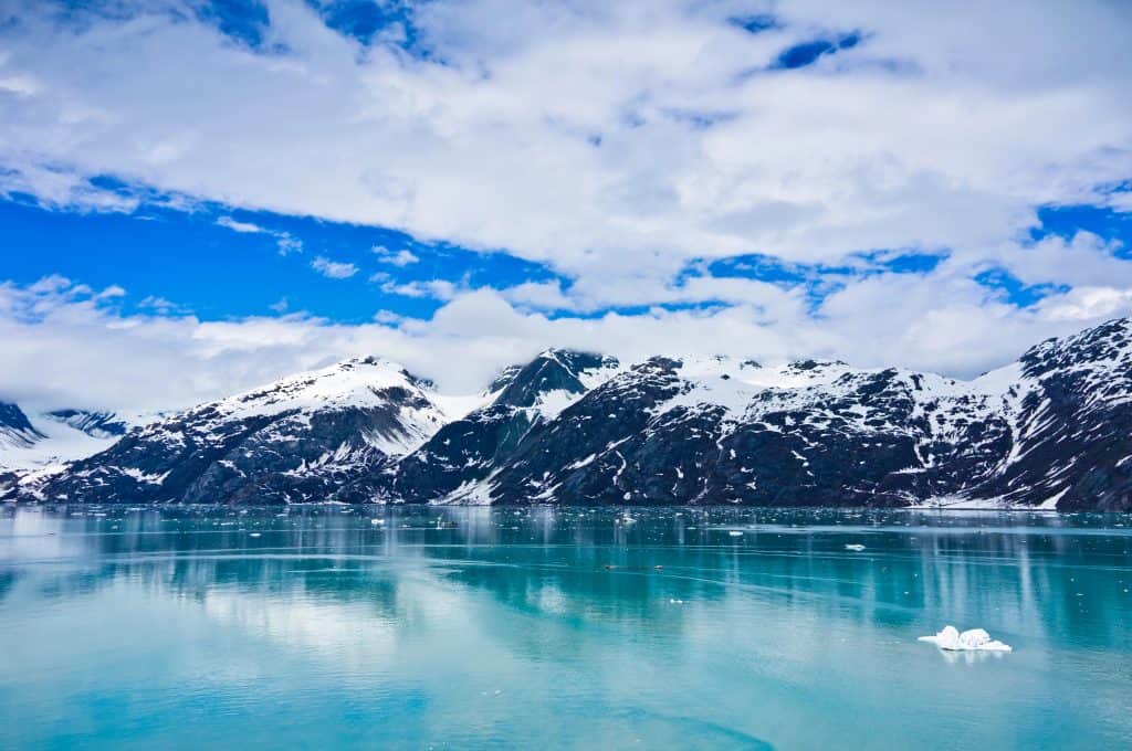 Experience the breathtaking beauty of Glacier Bay National Park, where snow-capped mountains and glaciers are reflected in the calm, turquoise water under a partly cloudy sky.