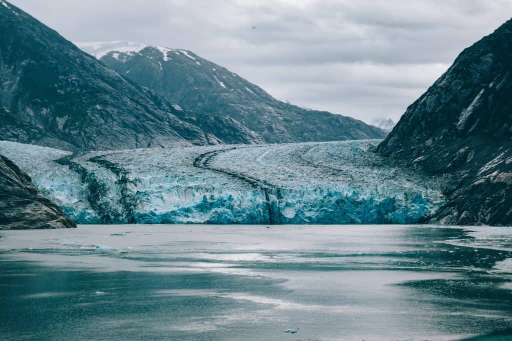 A glacier extends between two dark rocky mountains in Glacier Bay National Park, with icy blue tones and layered textures, reaching a calm, cold body of water under a cloudy sky. Experience one of the best things to do in Glacier Bay by exploring these breathtaking views.