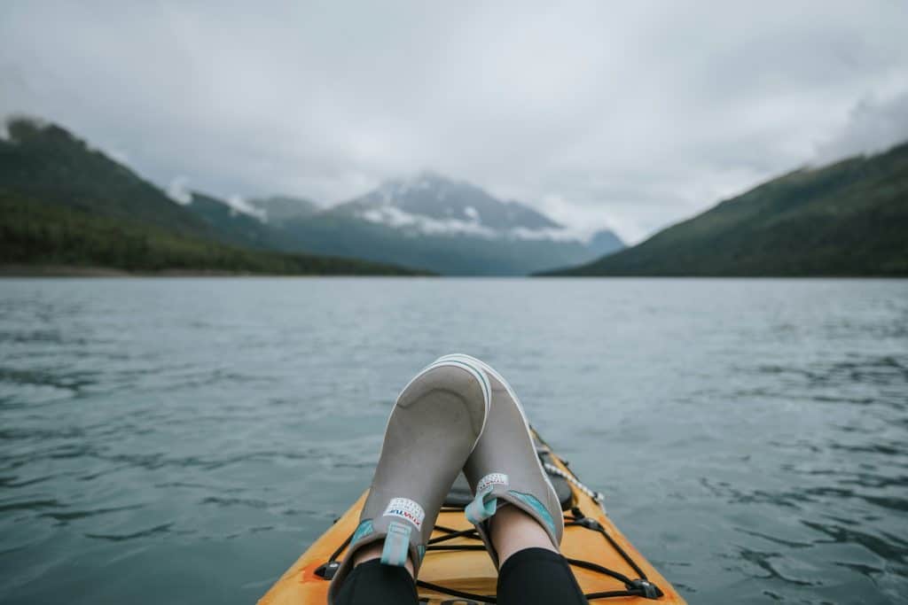 Relaxing with feet on a kayak, this person savors the serenity of a calm lake framed by misty mountains and an overcast sky, embodying the essence of Southeast Alaska outdoor activities.