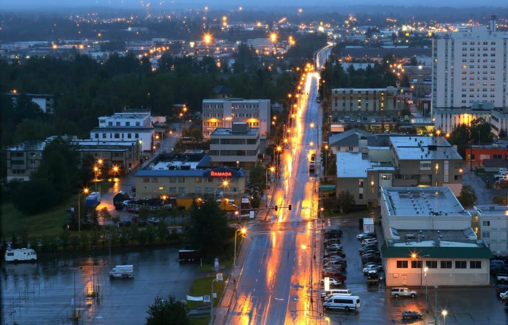 Aerial view of a cityscape at dusk showcases Anchorage attractions, with wet streets reflecting lights. Buildings line the long road while a cloudy Southcentral Alaska sky looms in the background.