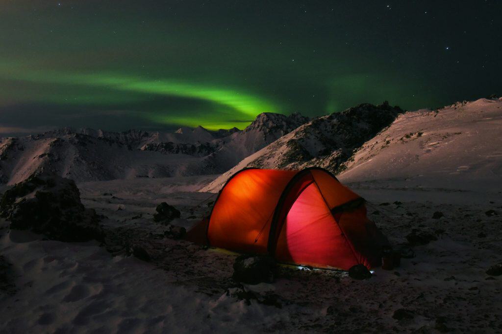 A glowing orange tent stands in the snowy expanse of Chugach State Park, set against a night sky alive with dancing green auroras—a true camping paradise for those seeking unforgettable outdoor adventures.