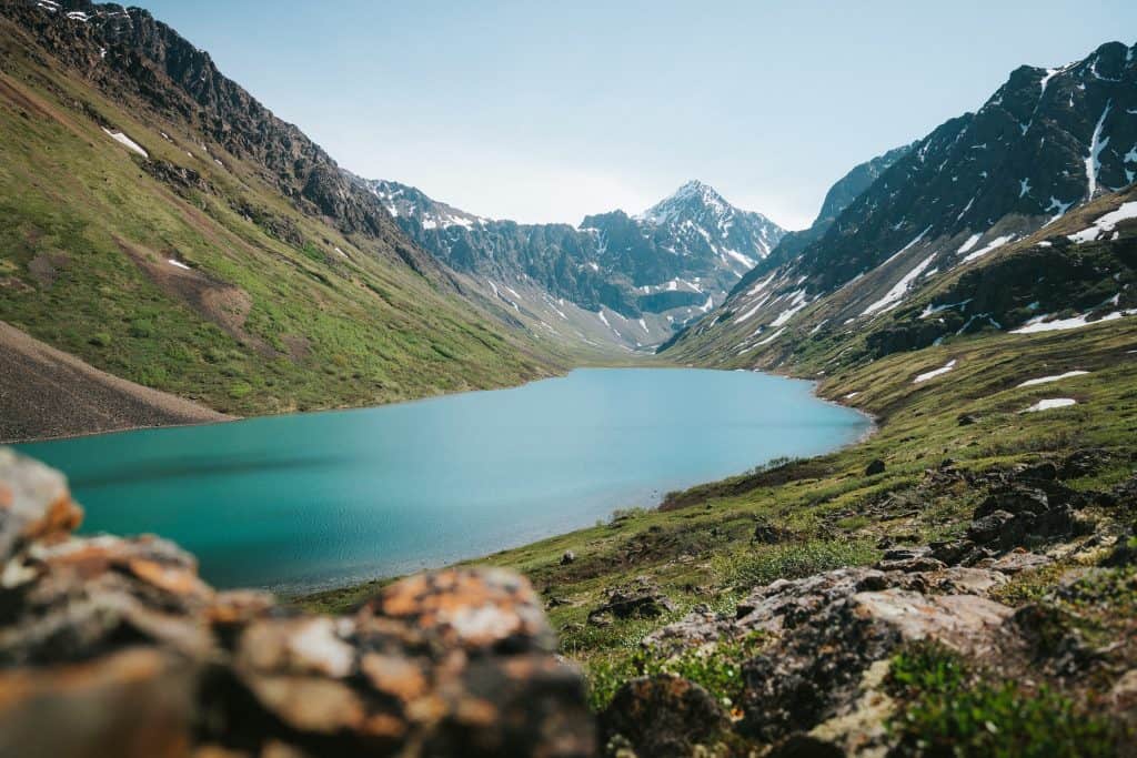 A serene mountain landscape with a turquoise lake surrounded by rocky peaks and patches of greenery under a clear blue sky, this scene captures the breathtaking beauty often found along the best hikes in Chugach State Park.