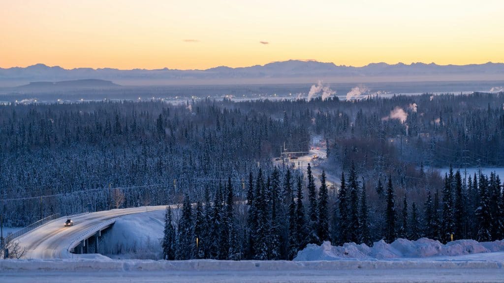 A snow-covered road winds through the forest with smoke rising from chimneys, set against a mountainous backdrop at dawn—a perfect scene for Interior Alaska travel enthusiasts.