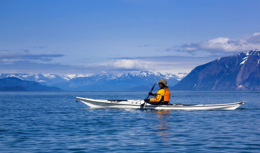 A person in a yellow jacket kayaks on calm, blue water in Glacier Bay, framed by snow-capped mountains under a clear sky, showcasing one of the best kayaking routes in Alaska.