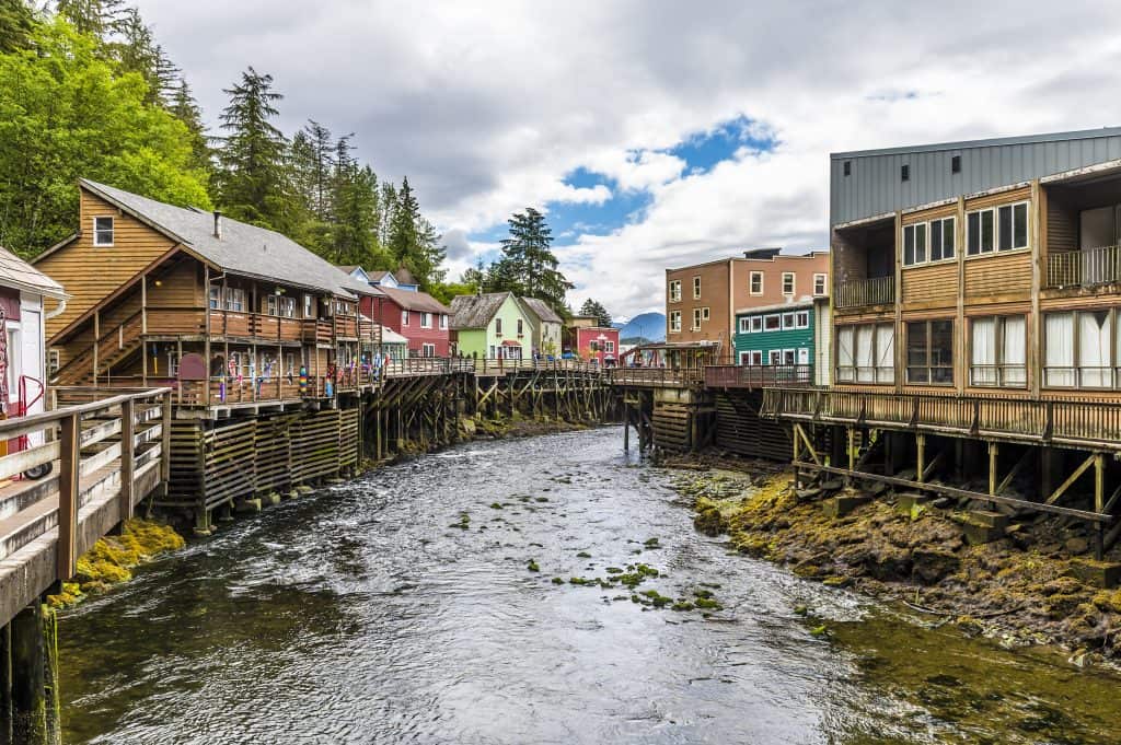 A narrow river flows between wooden buildings on stilts in a charming Alaskan village, with trees and a cloudy sky painting the backdrop of this delightful coastal town.