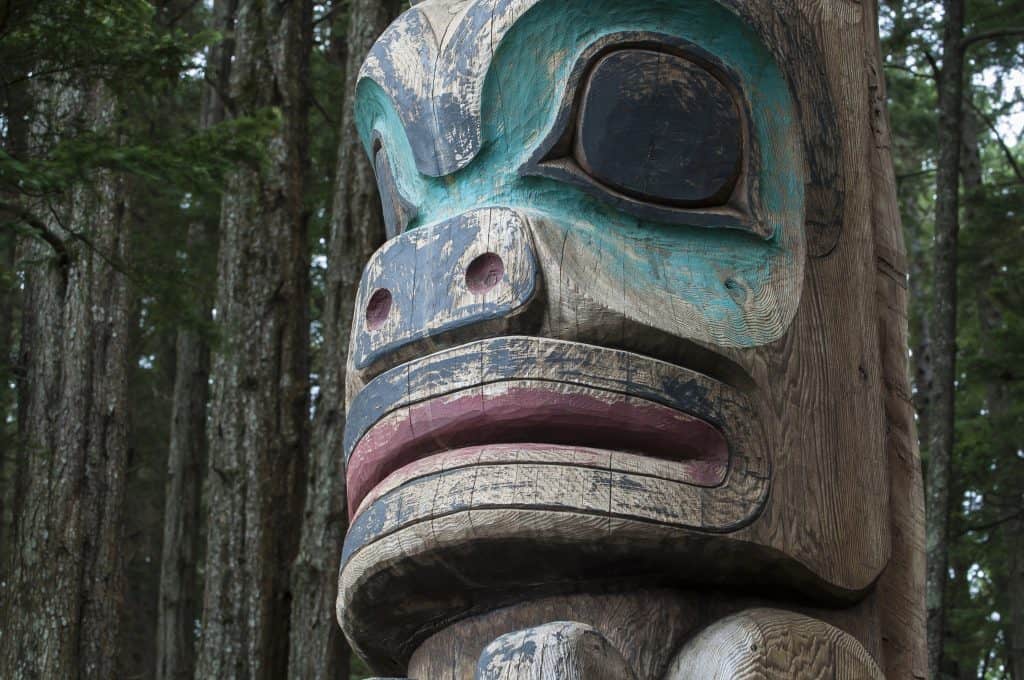 Close-up of a wooden totem pole with carved and painted details, reflecting Native Alaskan culture. It features large eyes and mouth, surrounded by trees in the background, offering a glimpse into the rich tapestry of Southeast Alaska history.