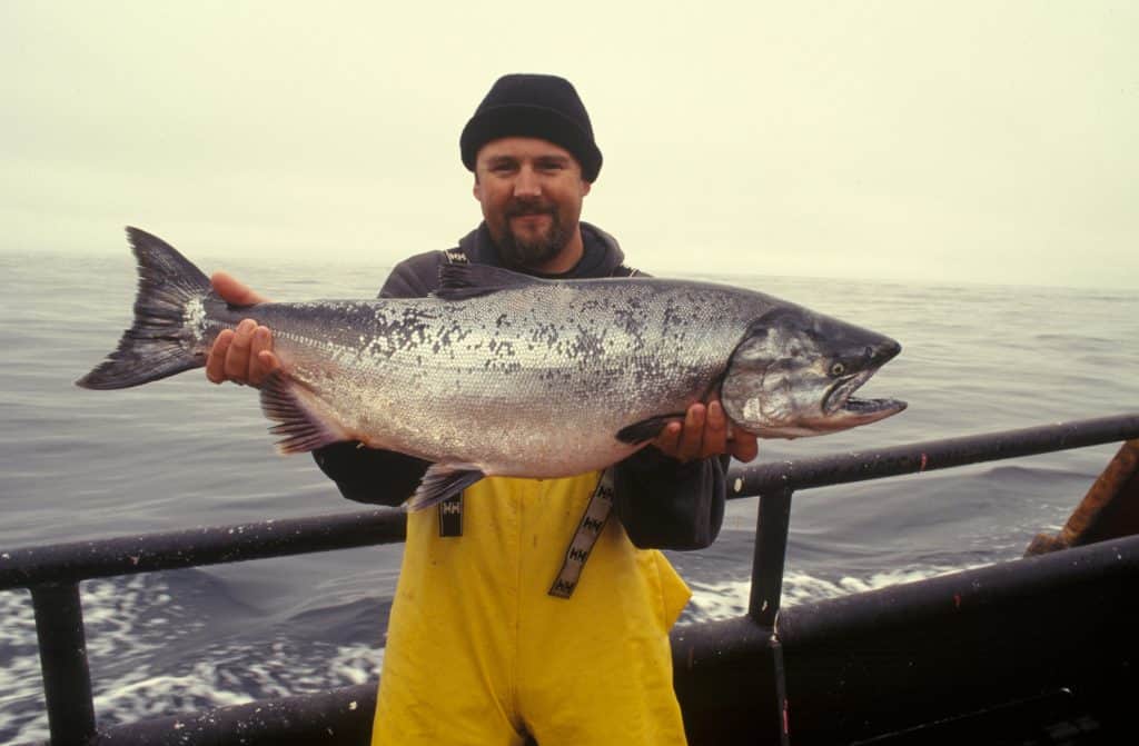 A person in yellow overalls and a black cap proudly holds a large fish on a boat, set against the stunning backdrop of the ocean. Discovering one of the best fishing spots in Southeast Alaska makes this catch even more exhilarating.