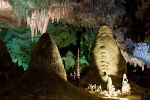 Large stalagmites and hanging stalactites inside a cavern, with textured rock formations illuminated by artificial lighting and a cool temp that adds to the mysterious atmosphere.
