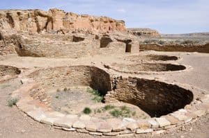 Ancient stone ruins with circular kivas set in a desert landscape, backed by rocky cliffs under a clear sky—explore the legacy of Chaco Culture with our 2025 Vacation Guide.