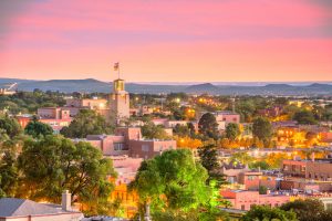 A cityscape view of Santa Fe, New Mexico, at sunset featuring adobe-style buildings, trees, and a skyline with distant mountains—perfect inspiration for your 2025 Vacation Guide.