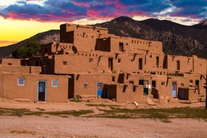 Adobe pueblo building with multiple stories and blue doors, set against mountains and a colorful sunset sky—a perfect scene for your Taos travel adventure.