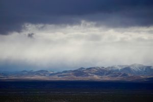 Dark clouds hang over distant sunlit mountains with patches of snow, while a flat, shadowed plain stretches across the foreground—a scene straight from a Truth or Consequences 2025 Vacation Guide.