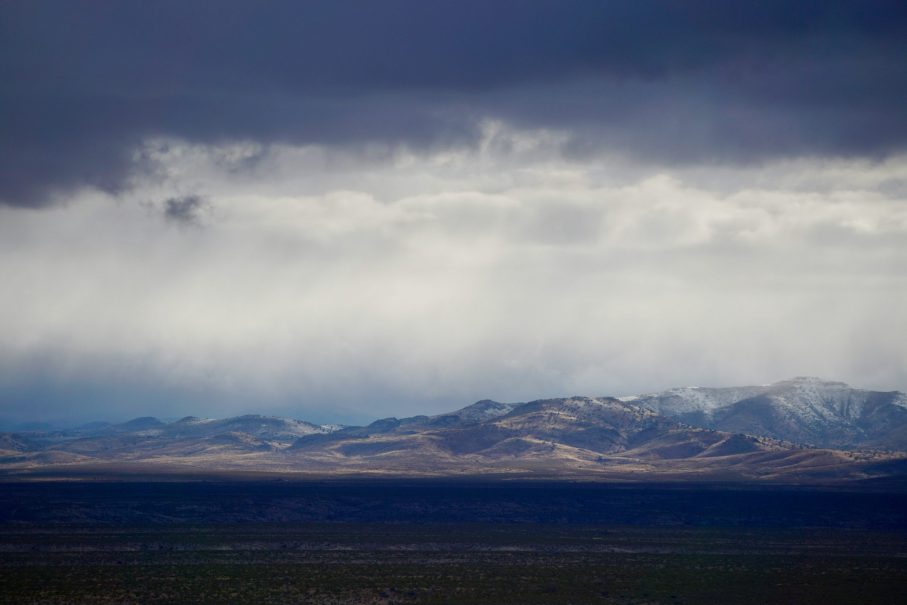Dark clouds hang over distant sunlit mountains with patches of snow, while a flat, shadowed plain stretches across the foreground—a scene straight from a Truth or Consequences 2025 Vacation Guide.