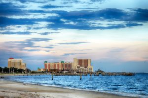 View of a sandy Biloxi beach with calm waves, wooden piers, and several hotels or buildings along the Mississippi shoreline under a partly cloudy sky, perfect for a relaxing Beach Getaway.