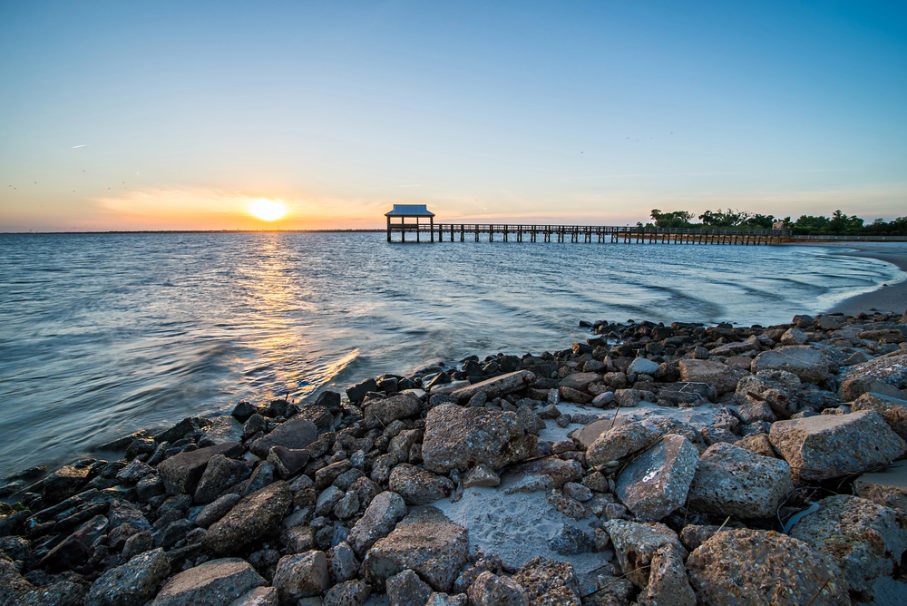 Rocky shore in the foreground, a long wooden pier stretching over Gulfport waters, and the sun setting on the horizon with a clear sky—perfect for your 2025 Vacation Guide.