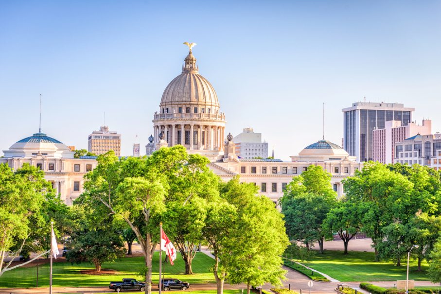 The Mississippi State Capitol building stands behind green trees in Jackson, with a clear sky and modern city buildings in the background.