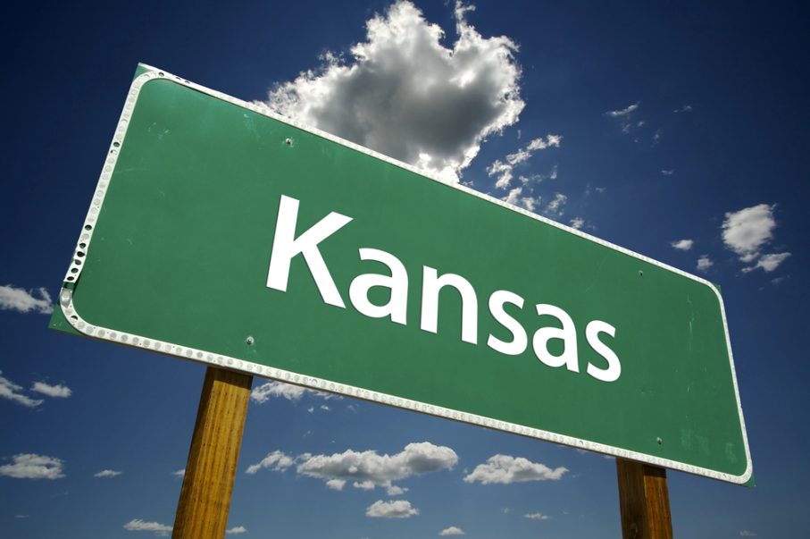 A green Kansas road sign with white text stands against a blue sky with scattered clouds, perfect for any KS travel guide.