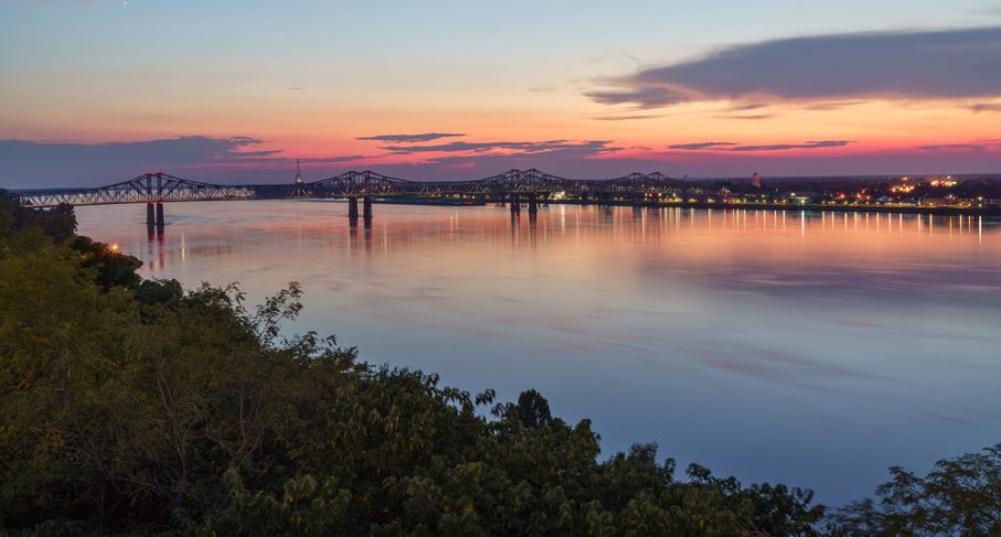 A long bridge spans a wide river at sunset in Natchez, with city lights and clouds visible in the distance and trees in the foreground—a scenic view perfect for any 2025 Vacation Guide.