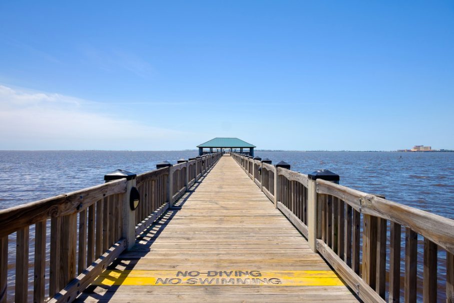 A wooden pier with railings extends over a body of water toward a covered structure; "No Diving No Swimming" and temp warnings are painted on the boards at the start of the pier.