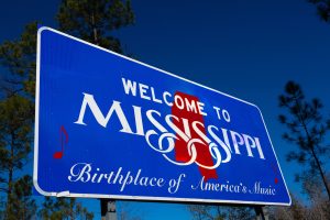 Blue "Welcome to Mississippi" road sign with red notes and text reading "Birthplace of America's Music," set against a clear blue sky and trees in the background—your perfect photo op on your Oxford travel adventure.