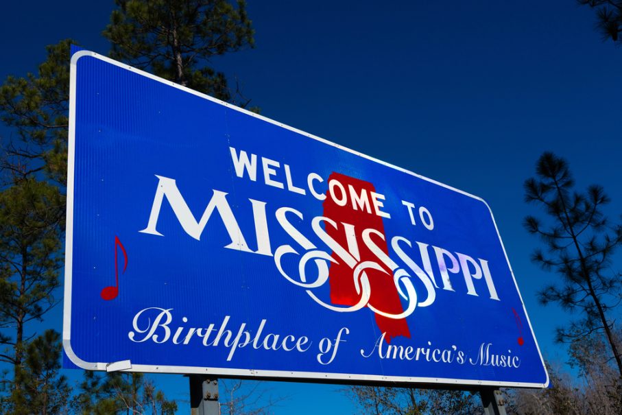 Blue "Welcome to Mississippi" road sign with red notes and text reading "Birthplace of America's Music," set against a clear blue sky and trees in the background—your perfect photo op on your Oxford travel adventure.