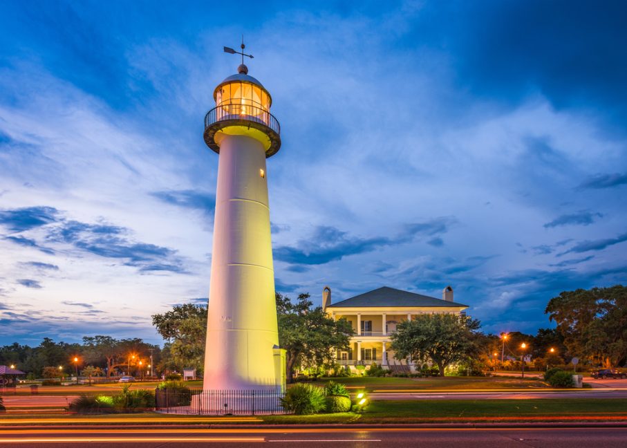 A tall white lighthouse stands near a large historic house at dusk, with streetlights and car light trails in the foreground—a classic scene among the best vacation spots in Mississippi.