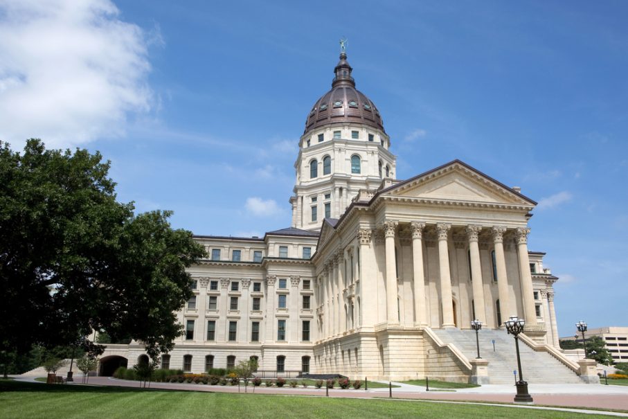 A neoclassical government building with a large dome, columns, and stone steps stands in Topeka, Kansas, set against a blue sky dotted with clouds and a tree in the foreground—a highlight for any Vacation Travel Guide.