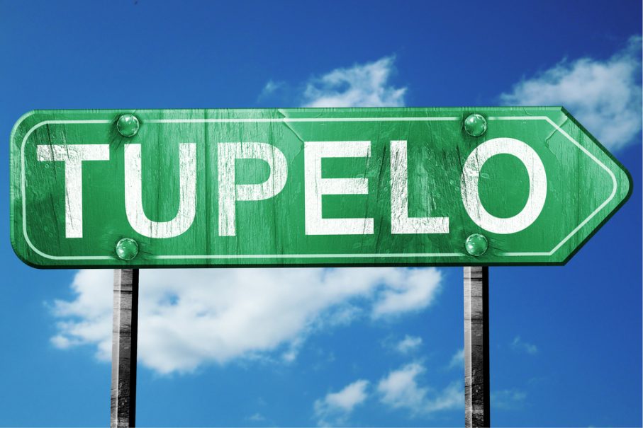 A green road sign with the word "TUPELO" in large white letters, set against a blue sky with scattered clouds—a classic sight for any Tupelo Travel guide.